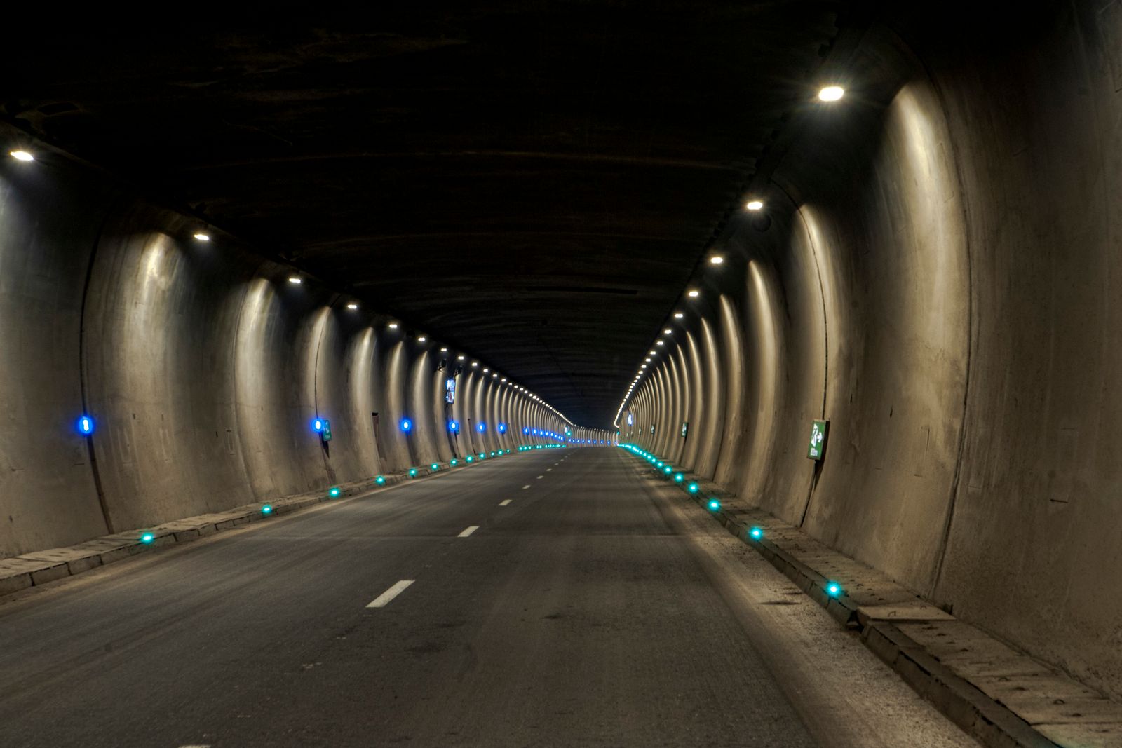 A dimly lit concrete road tunnel receding into darkness, blue marker lights running along both sides